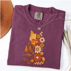 A folded purple t-shirt with a retro-style cat graphic made of orange, yellow, and white flowers and trees, styled on a white background with a necklace and a brown hat edge visible.