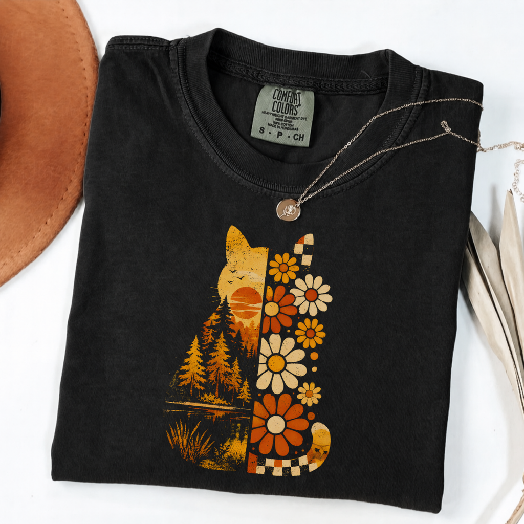 A folded black t-shirt with a retro-style graphic of a cat and flowers in orange, yellow, and white. The shirt is styled on a white background with a brown hat edge and a necklace visible.