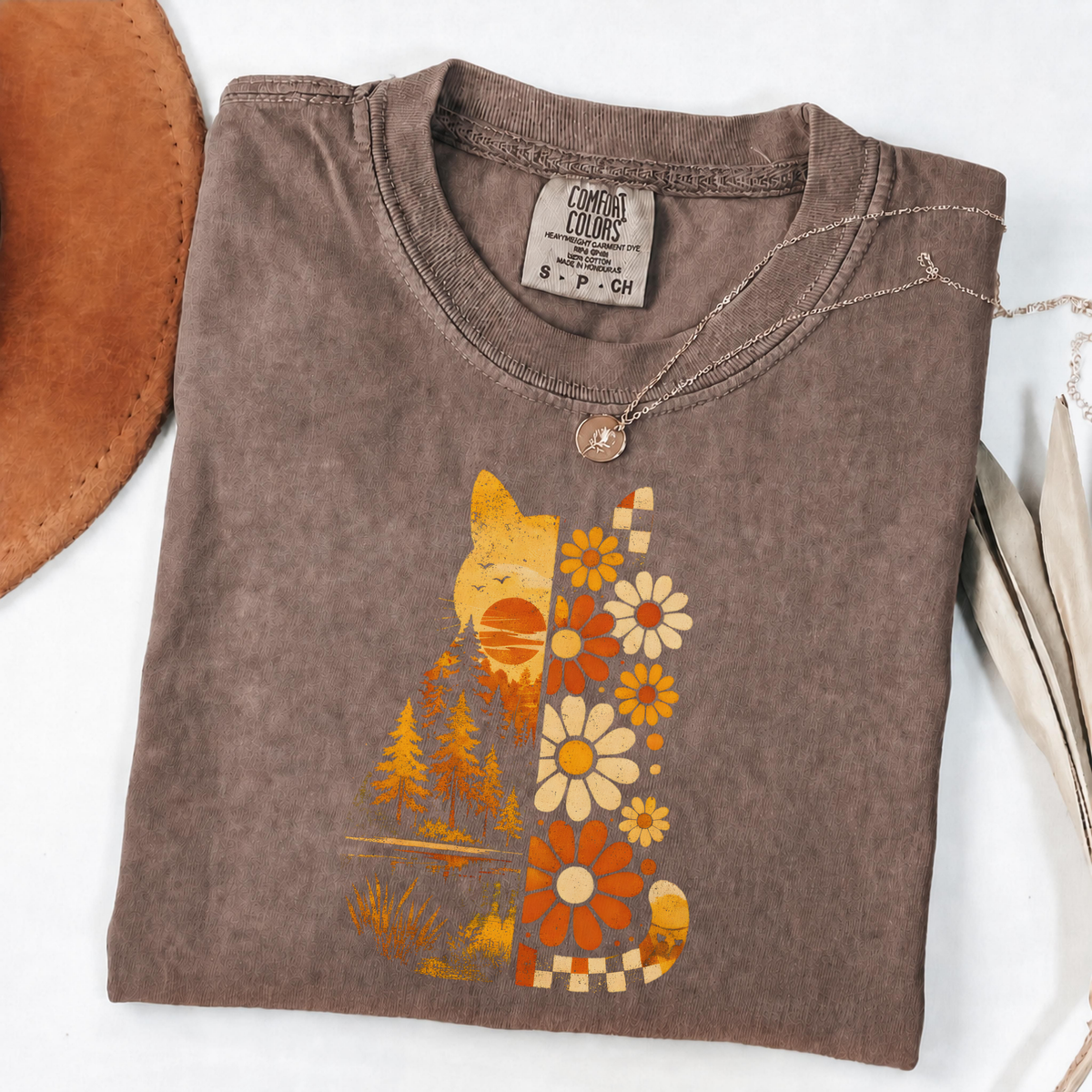 A folded brown t-shirt with a retro-style cat and floral graphic in orange, yellow, and cream, styled on a white background with a leather hat edge and a necklace visible.