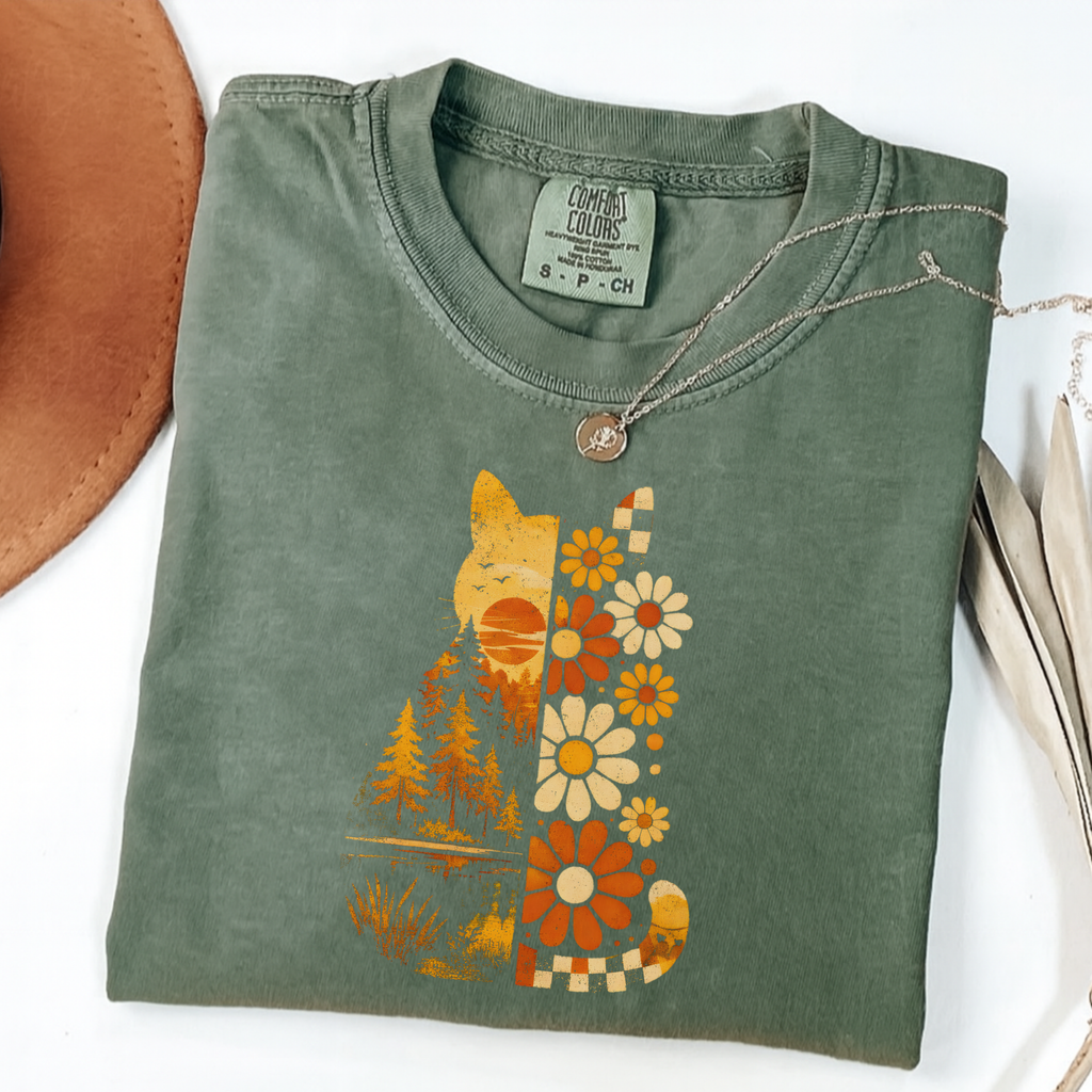 An olive green folded t-shirt with a retro-style graphic of a cat made of flowers, trees, and a sunset, styled on a white background with a brown hat and a necklace.