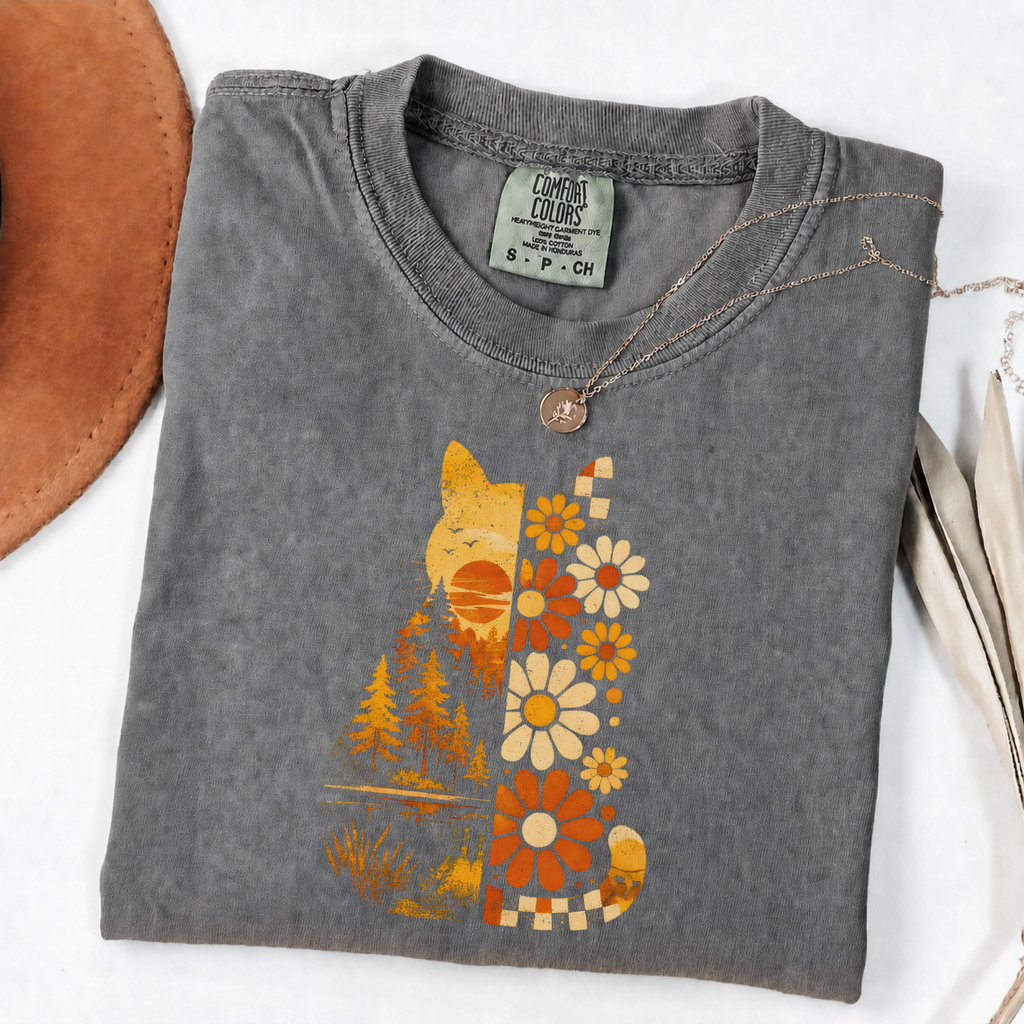 A folded gray t-shirt with a retro-style cat graphic made of yellow, orange, and white flowers and trees, styled on a white background with a brown leather hat edge and a necklace.