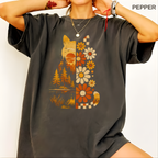 A woman wearing a dark charcoal gray oversized t-shirt with a split design: one side features a cat silhouette filled with trees and a lake landscape, while the other side features a pattern of orange, beige, and white flowers. The brand name 'PEPPER' is visible in the top right corner.